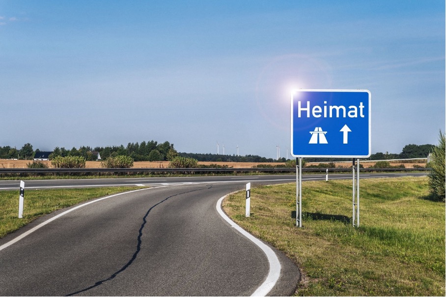 Ein Verkehrsschild mit der Aufschrift 'Heimat' zeigt in Richtung einer Straße, umgeben von einer ländlichen Landschaft mit blauem Himmel und Feldern im Hintergrund.